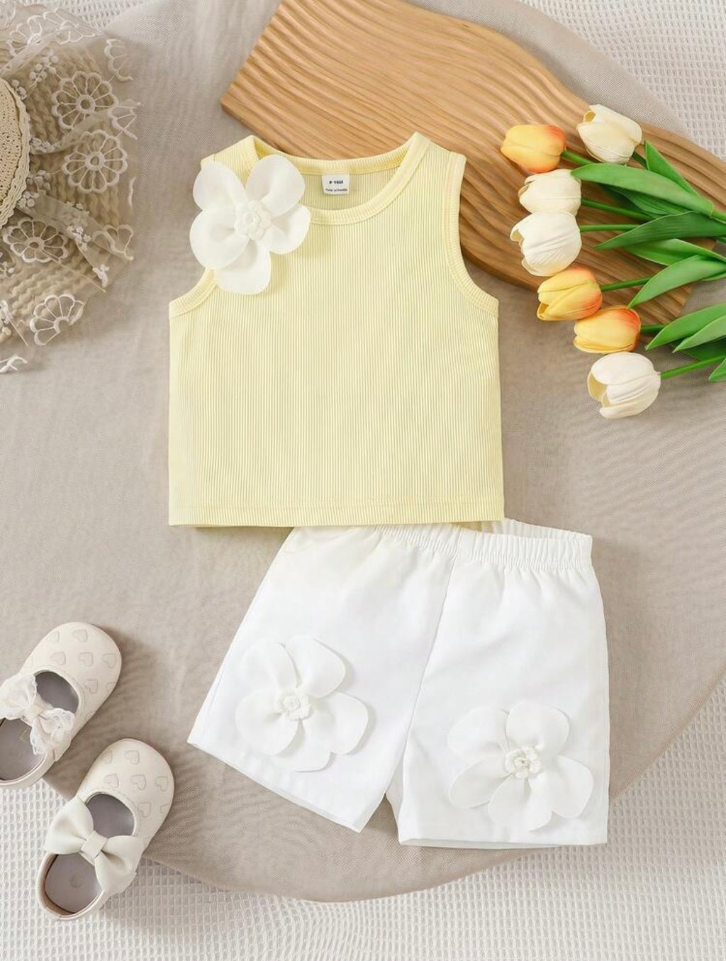 Children's outfit with yellow top and white shorts featuring flower details, accompanied by shoes and tulips on a neutral background.